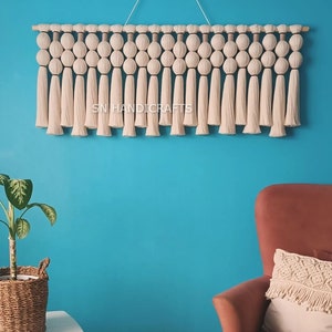 May include: A white macrame wall hanging with a wooden dowel and tassels. The hanging features a series of woven knots and is decorated with a small sign that reads "SN HANDICRAFTS".