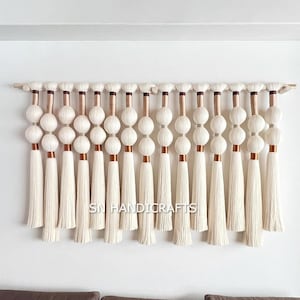 Op de afbeelding: Een crèmekleurige macramé wandhanger met een houten staaf. Het ontwerp heeft een reeks witte garen ballen en kwasten, geaccentueerd met koperkleurige metalen buizen. De tekst "SN HANDICRAFTS" is zichtbaar aan de onderkant.