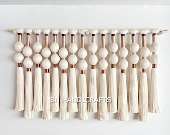 May include: A white macrame wall hanging with a wooden dowel at the top. The hanging features a series of white tassels with copper accents. The tassels are arranged in a row and are all the same length. The hanging is a great way to add a touch of bohemian style to any room.