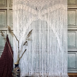 May include: A white macrame curtain with a geometric top design and long, vertical fringe. The curtain hangs in front of a wall with a decorative panel design. A vase with dried plants and a cat are in the foreground.