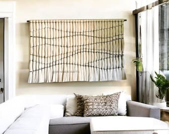 May include: A cream and grey woven wall hanging with a textured design, mounted on a wooden rod. The rectangular artwork hangs above a white sofa. The text "@SN HANDICRAFTS" is visible on the lower left of the wall hanging. The piece is a decorative item.