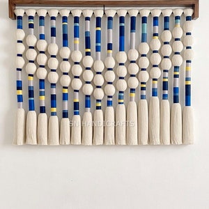 May include: A wall hanging made of white yarn with blue, yellow, and gray accents. The yarn is woven into a pattern of vertical stripes with small white balls spaced evenly throughout. The hanging is framed with a dark wood frame.