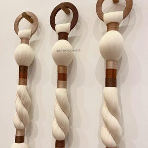 Könnte beinhalten: Drei dekorative Wandbehänge im natürlichen, böhmischen Stil. Jeder hat einen Holzring und -dübel, ein weißes, kugelförmiges Element und ein gedrehtes, cremefarbenes Seildesign. Die Aufhänger haben braune und beige Akzente und sind an einer weißen Wand befestigt.