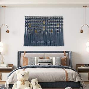 May include: A blue and gold wall hanging with tassels, featuring the text "SN HANDICRAFTS", above a bed with a white duvet and two teddy bears.