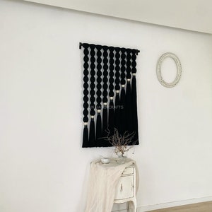 K&ouml;nnte beinhalten: Schwarze und wei&szlig;e Wandbeh&auml;nge aus Garn mit geometrischem Muster. Der Behang hat unten einen Fransen und ist an einer wei&szlig;en Wand befestigt.