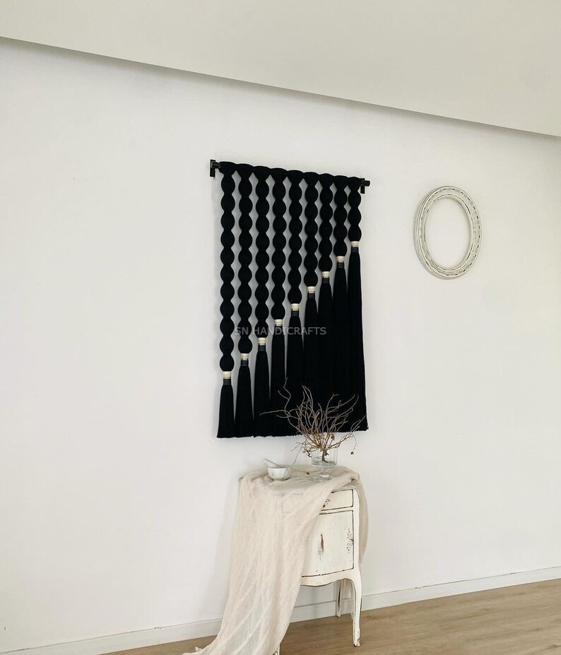 K&ouml;nnte beinhalten: Schwarze und wei&szlig;e Wandbeh&auml;nge aus Garn mit geometrischem Muster. Der Behang hat unten einen Fransen und ist an einer wei&szlig;en Wand befestigt.