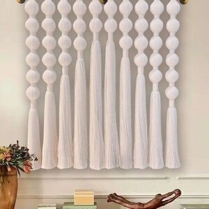 May include: White macrame wall hanging with a row of large white pom-poms and long white tassels.