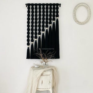 K&ouml;nnte beinhalten: Schwarze und goldene Makramee-Wandbeh&auml;nge mit geometrischem Design. Der Behang besteht aus einer Reihe schwarzer Garn-Schleifen mit goldenen Akzenten und langen schwarzen Quasten.