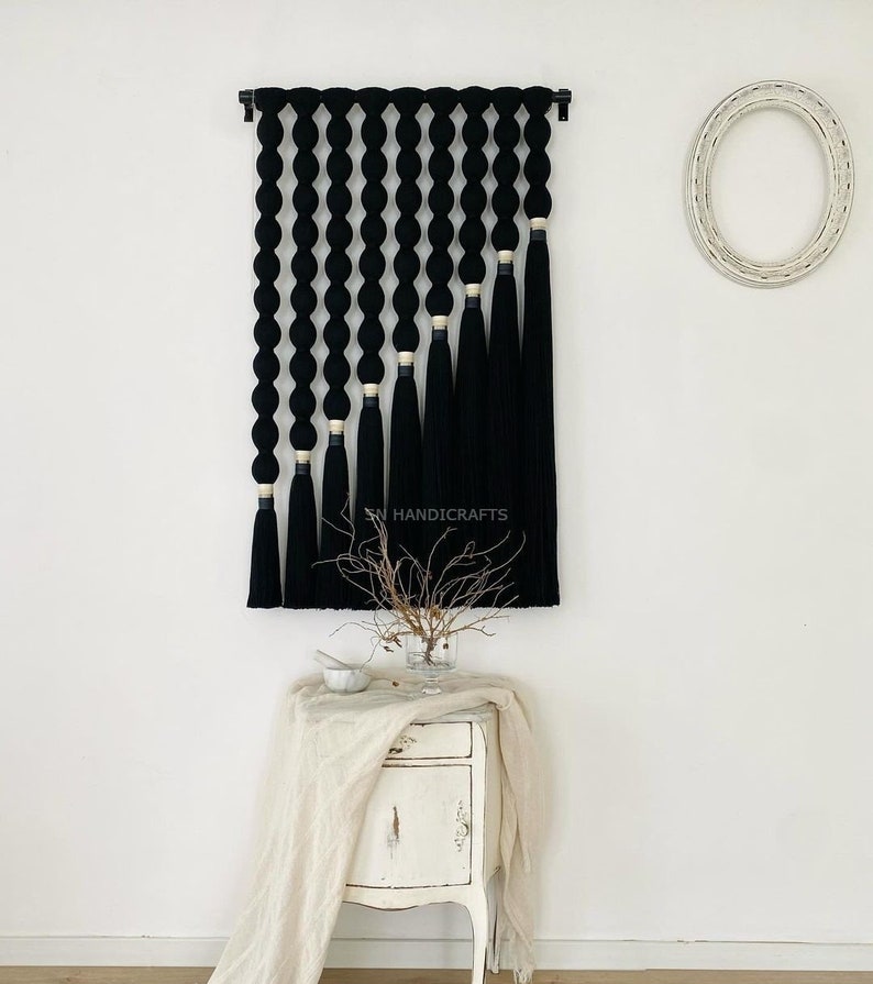 K&ouml;nnte beinhalten: Schwarze und goldene Makramee-Wandbeh&auml;nge mit geometrischem Design. Der Behang besteht aus einer Reihe schwarzer Garn-Schleifen mit goldenen Akzenten und langen schwarzen Quasten.