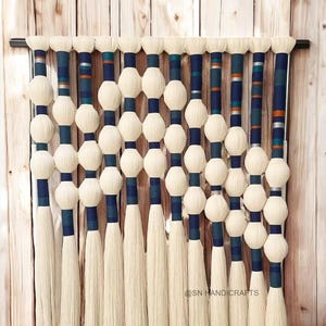 May include: A handcrafted wall hanging with cream-coloured spherical yarn ornaments, navy blue and teal accents, and long cream-coloured tassels. The design is mounted on a black rod against a wooden background. The piece is suitable for interior decor.