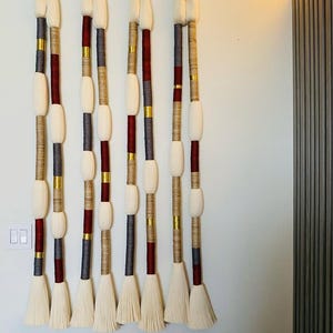 May include: Four vertical wall hangings with cream-colored tassels and oval accents. The hangings feature alternating sections of burgundy, gray, and gold-colored wrapped rope. The top of each hanging has a cream-colored, rounded shape.