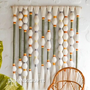 May include: A wall hanging made of white yarn with green, orange, and yellow accents. The hanging features a series of white pom-poms and is attached to a wooden dowel. The text "SN HANDICRAFTS" is visible at the bottom of the image.