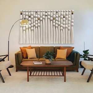 Puede incluir: Un tapiz de macram&eacute; blanco con detalles marrones y dorados. El tapiz presenta una serie de borlas y nudos, creando un dise&ntilde;o texturizado y moderno. El tapiz se muestra en un ambiente de sala de estar, sobre un sof&aacute; marr&oacute;n con almohadas y una mesa de caf&eacute;.