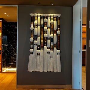May include: A large, vertical wall hanging with a series of cylindrical and spherical elements in shades of white, brown, and beige. The design includes long, white tassels at the bottom. The artwork is mounted on a dark gray wall.