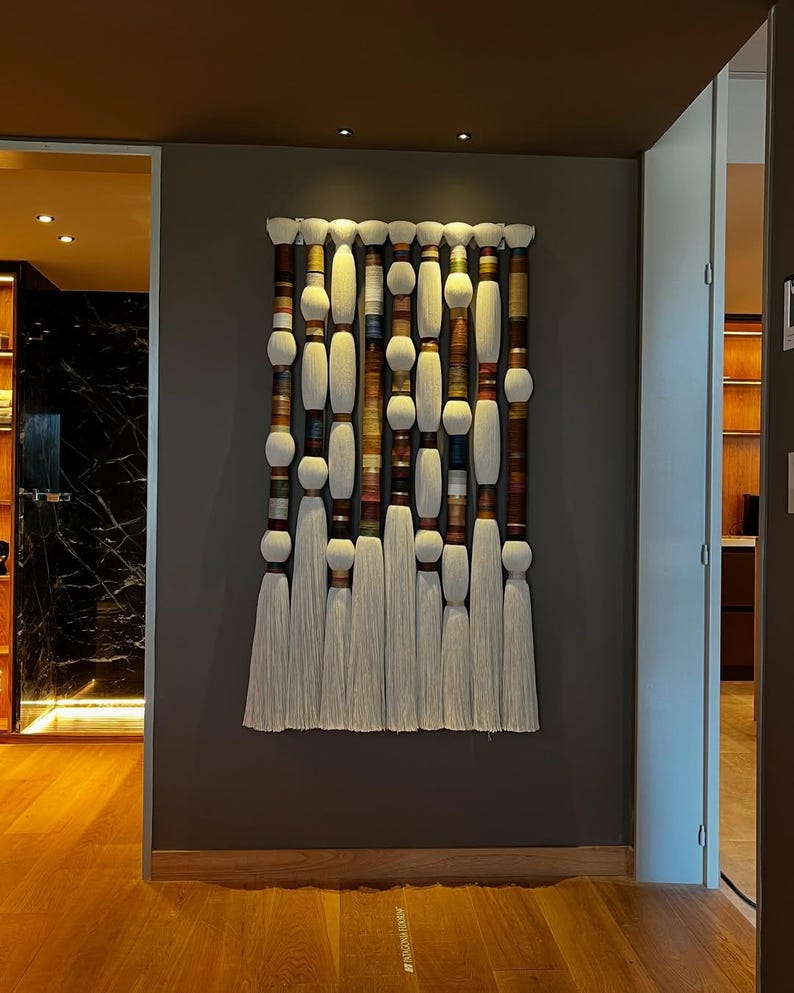 May include: A large, vertical wall hanging with a series of cylindrical and spherical elements in shades of white, brown, and beige. The design includes long, white tassels at the bottom. The artwork is mounted on a dark gray wall.