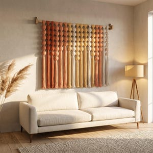 Könnte beinhalten: Ein dekorativer Wandbehang mit Farbverlauf von Orange nach Gelb, bestehend aus runden, strukturierten Elementen und langen Quasten. Darunter befindet sich ein cremefarbenes Sofa. Der Wandbehang ist etwa 150 cm breit.