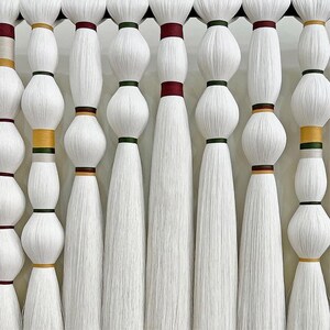 Puede incluir: Primer plano de diez hebras de hilo blanco, cada una con una banda de color diferente en la parte superior. Las hebras cuelgan de una barra de madera.