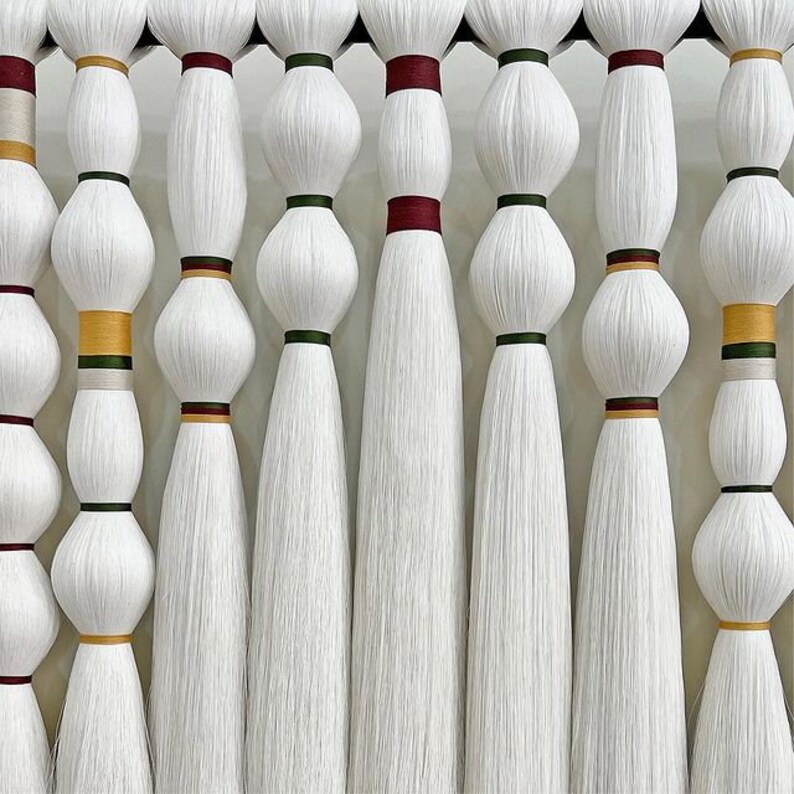 Puede incluir: Primer plano de diez hebras de hilo blanco, cada una con una banda de color diferente en la parte superior. Las hebras cuelgan de una barra de madera.