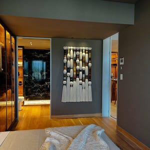 May include: A decorative wall hanging with a rectangular design. It features a series of white, brown, and cream-colored cylindrical and oval shapes, with white tassels hanging from the bottom. The artwork is mounted on a gray wall in a bedroom setting.