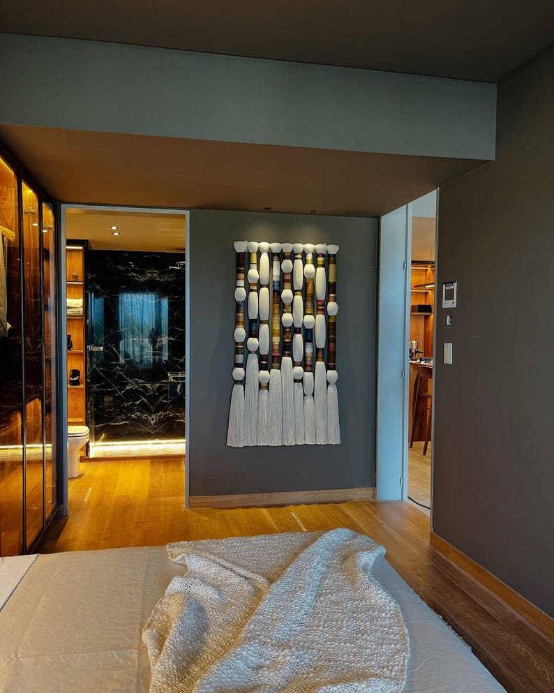 May include: A decorative wall hanging with a rectangular design. It features a series of white, brown, and cream-colored cylindrical and oval shapes, with white tassels hanging from the bottom. The artwork is mounted on a gray wall in a bedroom setting.