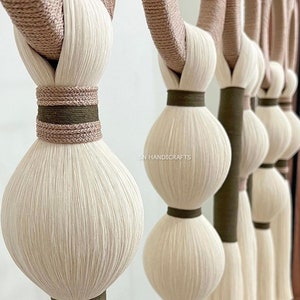 Puede incluir: Un tapiz de macram&eacute; hecho con m&uacute;ltiples hilos de lana blanca y beige, anudados y tejidos juntos. El tapiz presenta una serie de bolas redondas y esponjosas de lana, con un acento marr&oacute;n y beige.