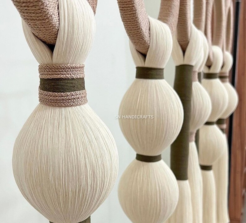 Puede incluir: Un tapiz de macram&eacute; hecho con m&uacute;ltiples hilos de lana blanca y beige, anudados y tejidos juntos. El tapiz presenta una serie de bolas redondas y esponjosas de lana, con un acento marr&oacute;n y beige.