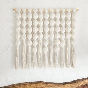 May include: Cream-colored macrame wall hanging featuring a series of large, spherical knots suspended from a natural wooden dowel. Long, textured tassels hang below the knots, adding a bohemian touch to the decor. This piece is ideal for adding texture and visual interest to any space.