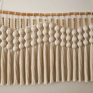 May include: A macrame wall hanging with white tassels and wooden beads. The hanging features a series of knotted balls and is attached to a wooden dowel.