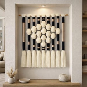 May include: A woven wall hanging with a geometric design. The piece features alternating black and beige vertical sections, with cream-colored yarn spheres and tassels. The wall art is mounted on a wooden rod, creating a modern, textured aesthetic.