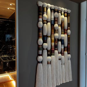 May include: A large, rectangular wall hanging featuring multiple vertical strands. Each strand has a white, fluffy top and bottom, with a central section wrapped in colorful threads. The piece is mounted on a gray wall.