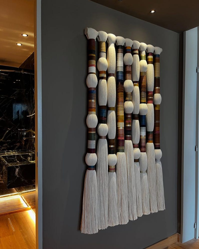 May include: A large, rectangular wall hanging featuring multiple vertical strands. Each strand has a white, fluffy top and bottom, with a central section wrapped in colorful threads. The piece is mounted on a gray wall.
