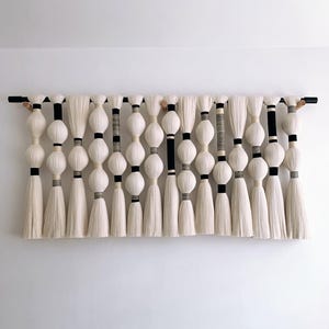 May include: A large, off-white fiber art wall hanging. The piece features a series of large, round, off-white shapes interspersed with black and gray accents, and long, white tassels. The artwork is mounted on a black wooden rod.
