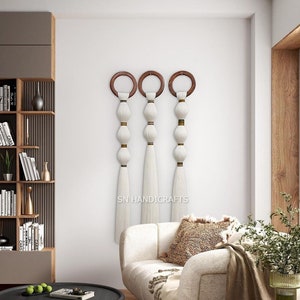 Puede incluir: Tres tapices de macramé blancos con anillos de madera y cuentas doradas. Los tapices se muestran en una pared blanca en un ambiente de sala de estar con un sofá blanco y una planta verde.