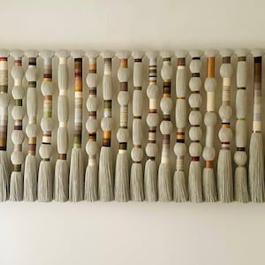 May include: A large, textured wall hanging featuring multiple vertical strands. Each strand has a light gray tassel at the bottom, with a series of wrapped sections in varying colors and textures, including beige, brown, and green. The background is a plain white wall.