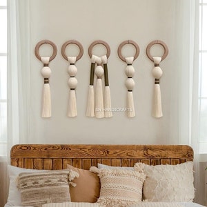 Puede incluir: Cinco tapices de macram&eacute; de pared con anillos de madera y borlas. Los tapices son en tonos marrones, beige y verdes. Los tapices est&aacute;n dispuestos en una fila en una pared blanca. El texto "SN HANDICRAFTS" es visible debajo de los tapices.