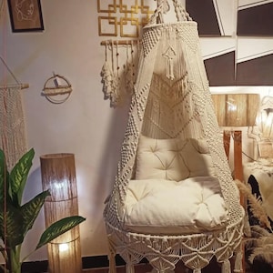 May include: Cream-colored macrame hanging chair with a tufted cushion and decorative fringe. The chair is suspended from the ceiling and features intricate knotwork. The chair is part of a bohemian-style interior.