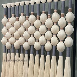 May include: A wall hanging made of white yarn with gray and brown accents. The yarn is woven into a series of round shapes, creating a textured and modern design. The hanging is attached to a black metal frame.