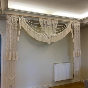 May include: A large, white macrame wall hanging with a decorative, arched design. The hanging is made of thick cotton cord and features a variety of knots and tassels.