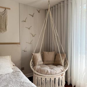 May include: A cream-coloured hanging chair with a round seat and back, featuring a macrame fringe. The chair is suspended from the ceiling and has beige cushions. The wall behind the chair has decorative bird accents.