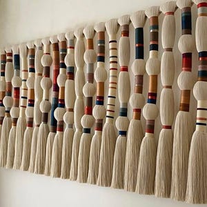 May include: A large, textured wall hanging with multiple vertical strands. Each strand features a series of cream-colored, rounded shapes and tassels. The strands are accented with bands of color, including red, blue, and brown.