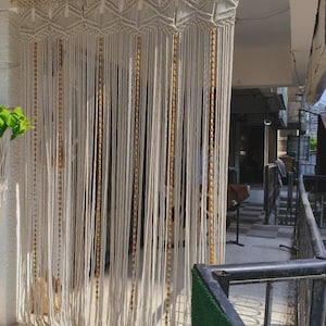 Può includere: Una tenda in macramè bianco sporco con un motivo geometrico nella parte superiore e lunghi fili verticali. La tenda presenta fili di perline color oro ed è appesa a un'asta di legno.