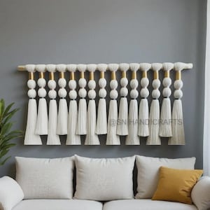 Könnte beinhalten: Weißer Makramee-Wandbehang mit Holzstab und goldenen Akzenten. Das Design zeigt mehrere Reihen weißer Quasten und kugelförmiger Perlen, die eine strukturierte, böhmische Ästhetik erzeugen. Wanddekoration über einem weißen Sofa.