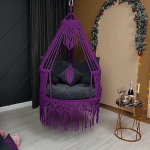 May include: A vibrant purple macrame hanging chair featuring intricate knotted patterns, a conical top structure, and long decorative fringe along the base. The chair includes a dark gray circular seat cushion and a matching square throw pillow.