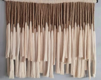 May include: A wall hanging made of brown and white yarn with a fringe. The yarn is woven together to create a textured look. The fringe is long and flowing.