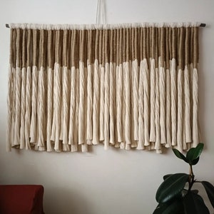 Op de afbeelding: Een wanddecoratie gemaakt van natuurlijke vezels met een bruin en wit kleurenschema. De decoratie is gemaakt van een reeks lange strengen garen die aan de bovenkant aan elkaar zijn gebonden. De strengen zijn in een verspringend patroon gerangschikt, waardoor een textuur en een visueel interessant ontwerp ontstaat.
