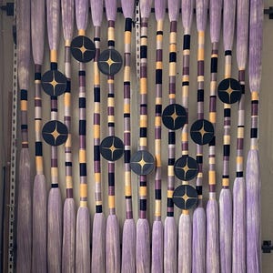 May include: A decorative wall hanging featuring multiple vertical elements. Each element has a lavender-colored, brush-like top and a striped body in shades of purple, black, and yellow. Dark gray circular accents with gold starburst designs are interspersed throughout.