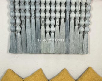 May include: A decorative wall hanging with a gradient of gray tassels and beaded accents. The text "SN HANDICRAFTS" is visible. Below, four square yellow throw pillows rest on a yellow surface, adding a pop of color.