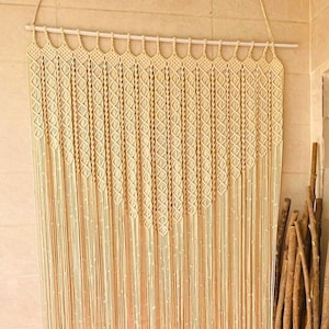 May include: A cream-colored macrame curtain with a diamond pattern hangs from a white rod. The curtain is made of thick cotton cord and has a fringe at the bottom.