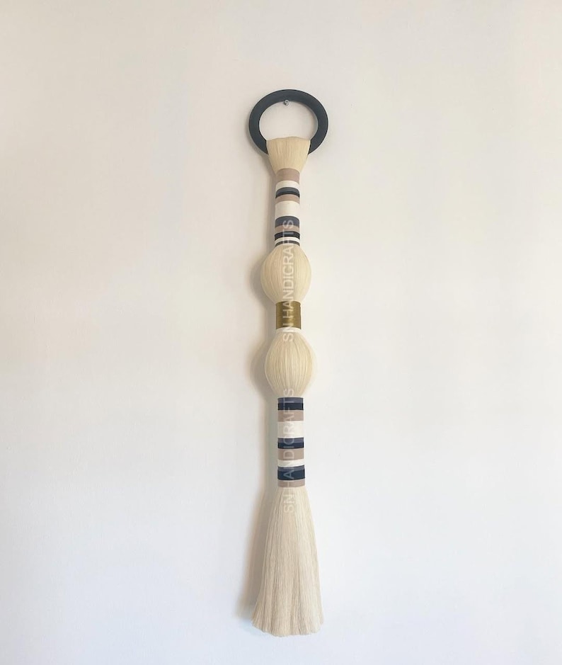 Puede incluir: Un tapiz de macram&eacute; blanco para colgar en la pared con rayas negras y beige y tres cuentas doradas y blancas. El tapiz est&aacute; sujeto a un anillo negro.