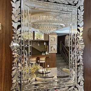 Può includere: Specchio rettangolare con una cornice in vetro incisa e ornata. La cornice presenta un design a foglie e fiori. Lo specchio riflette una stanza con un lampadario, un tavolo da pranzo e una scala. Lo specchio è montato su una parete di legno scuro.